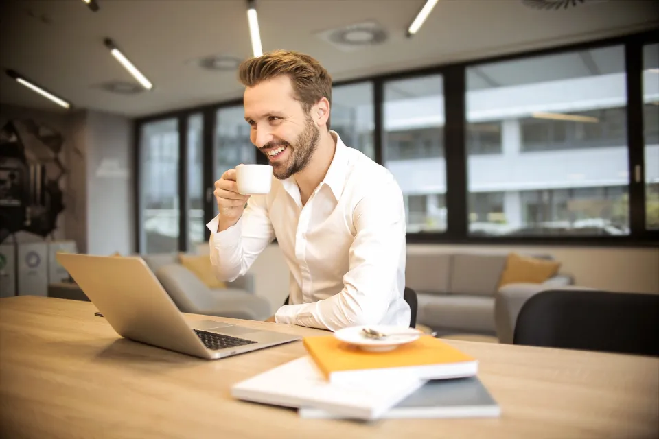 A smiling man with a coffee working on a laptop in a modern office