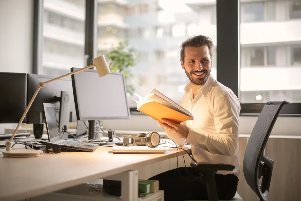 A man reading a printed report at a desk with monitors in a bright office