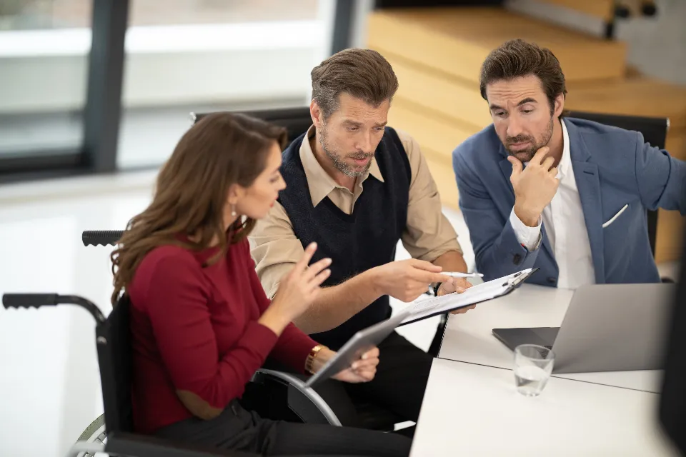Three colleagues at a meeting table reviewing a plan on a clipboard together