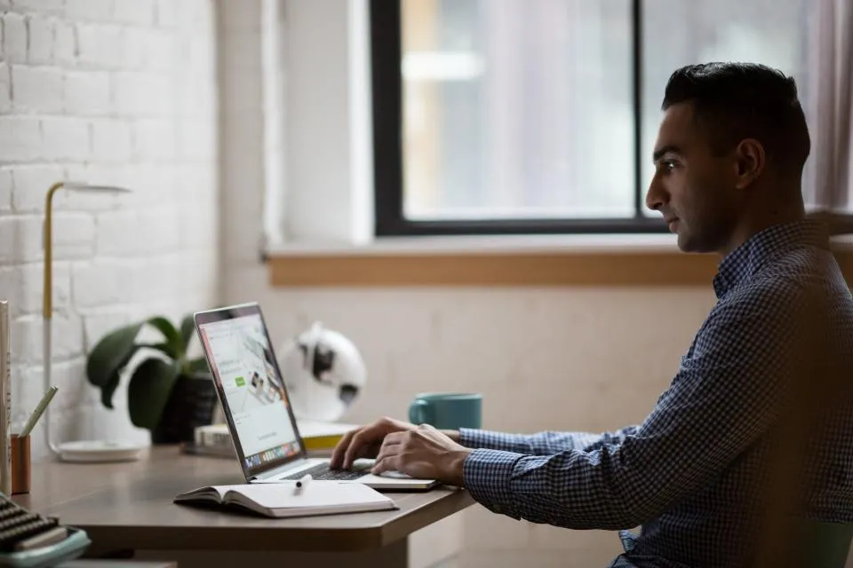 A man working on a laptop at a desk beside a large window